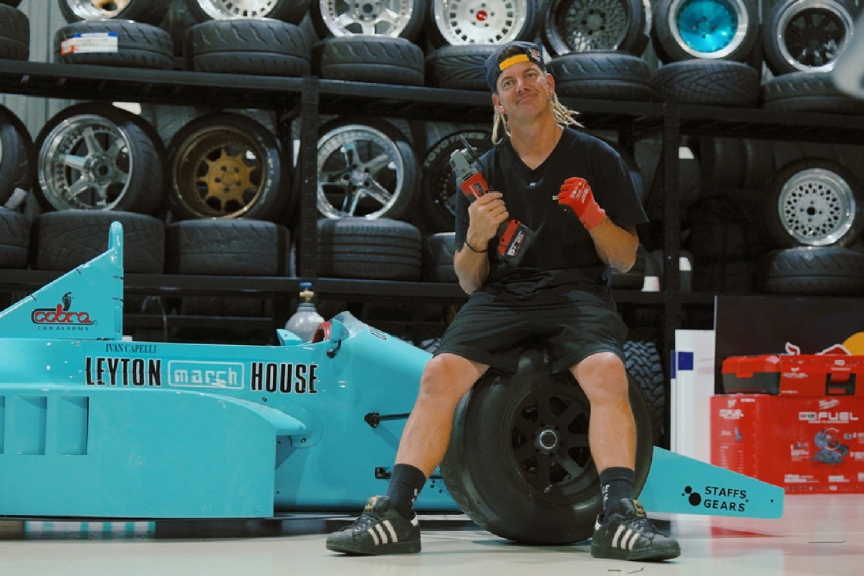 Builder sitting beside a teal Leyton House March Formula One chassis inside a workshop during the FORMIDABUL drift car project.